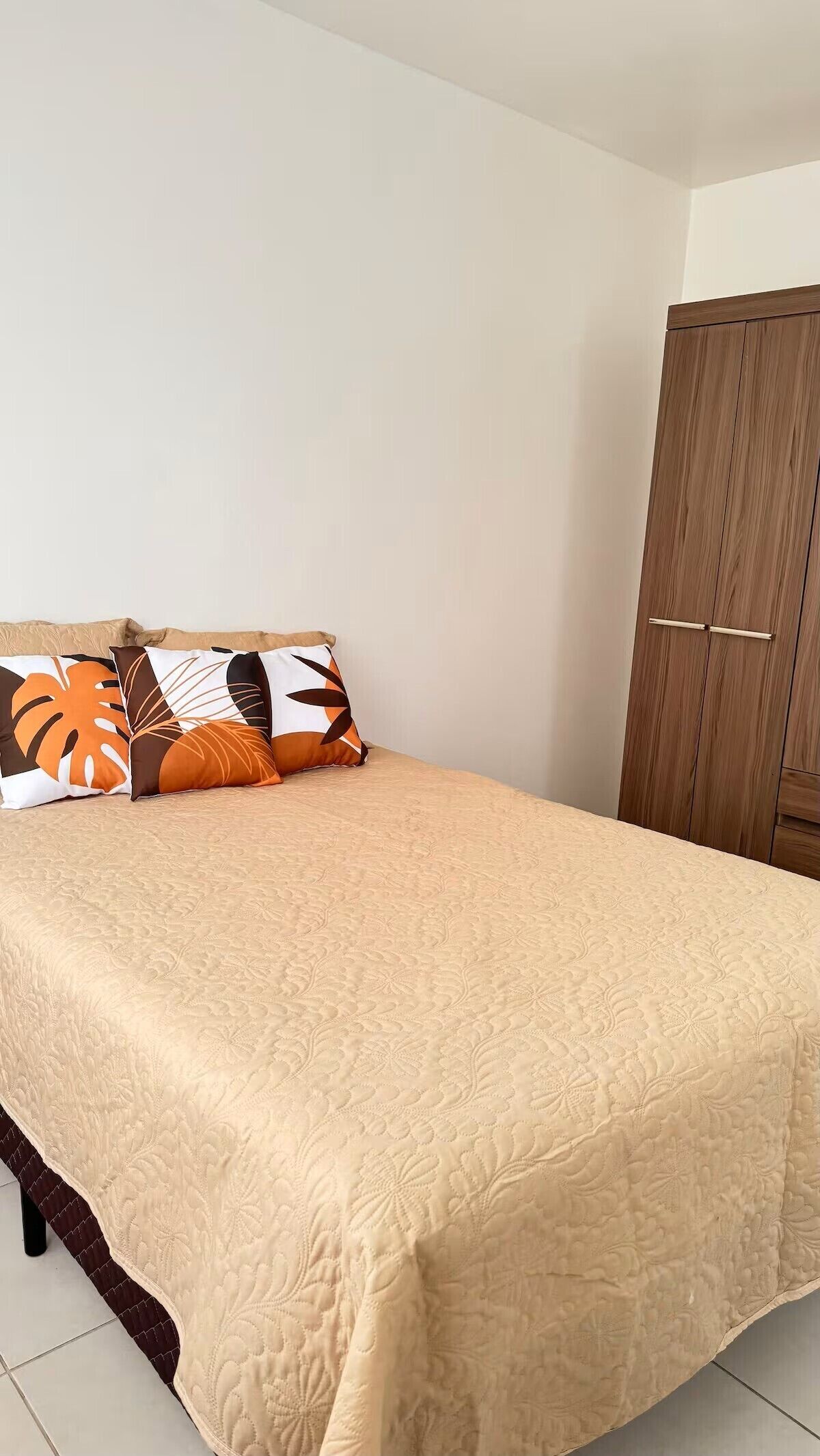 A bedroom with a bed, two decorative pillows, a wooden wardrobe, and a tiled floor.