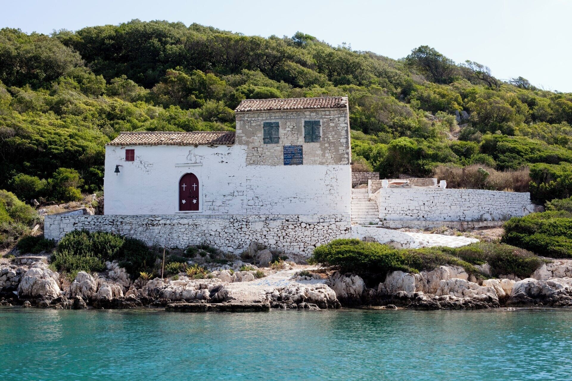 A white building with a red door and a small tower, surrounded by rocky terrain and greenery.