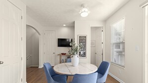 Un coin repas avec une table ronde, quatre chaises bleues, un vase blanc et un téléviseur fixé au mur.