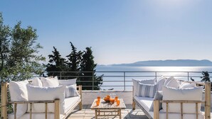 Une terrasse avec deux canapés blancs dotés de coussins moelleux et une table où sont disposées des boissons, offrant une vue sur une étendue d’eau et des montagnes lointaines.
