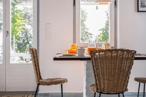 Ein Essbereich mit Korbstühlen, einem Tisch mit einer Karaffe Orangensaft und Blick auf Grünflächen außerhalb.