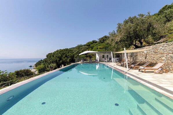 Una piscina rettangolare con acqua cristallina, circondata da un muro in pietra e un solarium in legno con lettini.