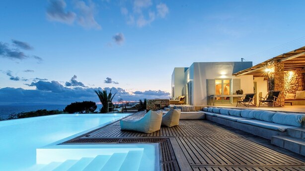 A modern outdoor pool area with a wooden deck, lounge seating, and a view of the sea. - Laeticia - Seven Bedroom Villa, Sleeps 14 (Finikas)