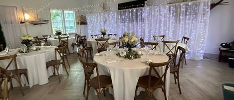 Un salón amplio con mesas redondas, sillas de madera y luces tipo guirnalda.