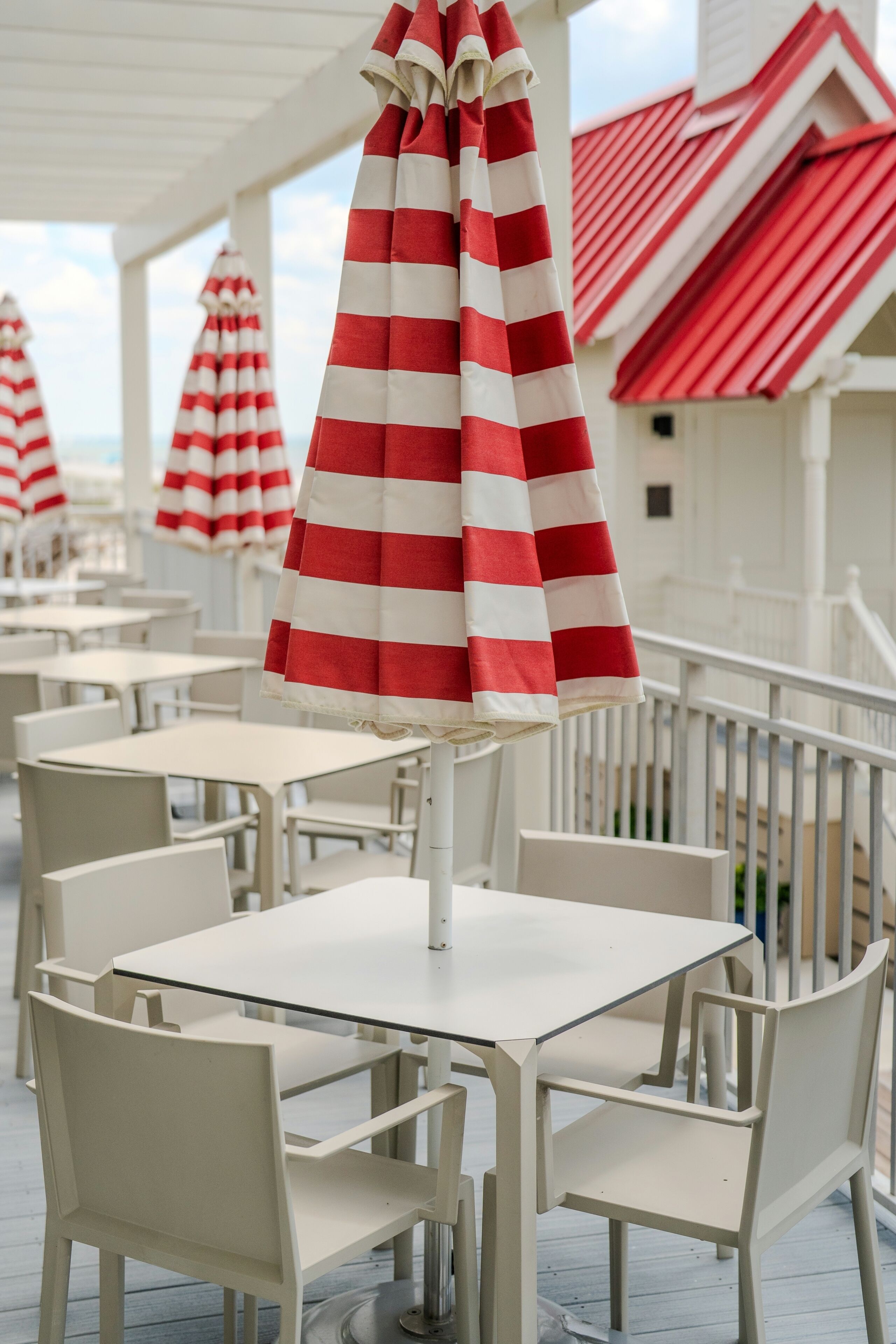 A covered outdoor seating area with tables and chairs, red and white striped umbrellas, and a building with a red roof.