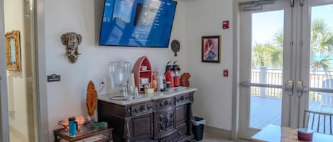 A room with a TV mounted on the wall, a wooden cabinet with various items, a glass coffee table, and a view through a sliding glass door to an outdoor area.