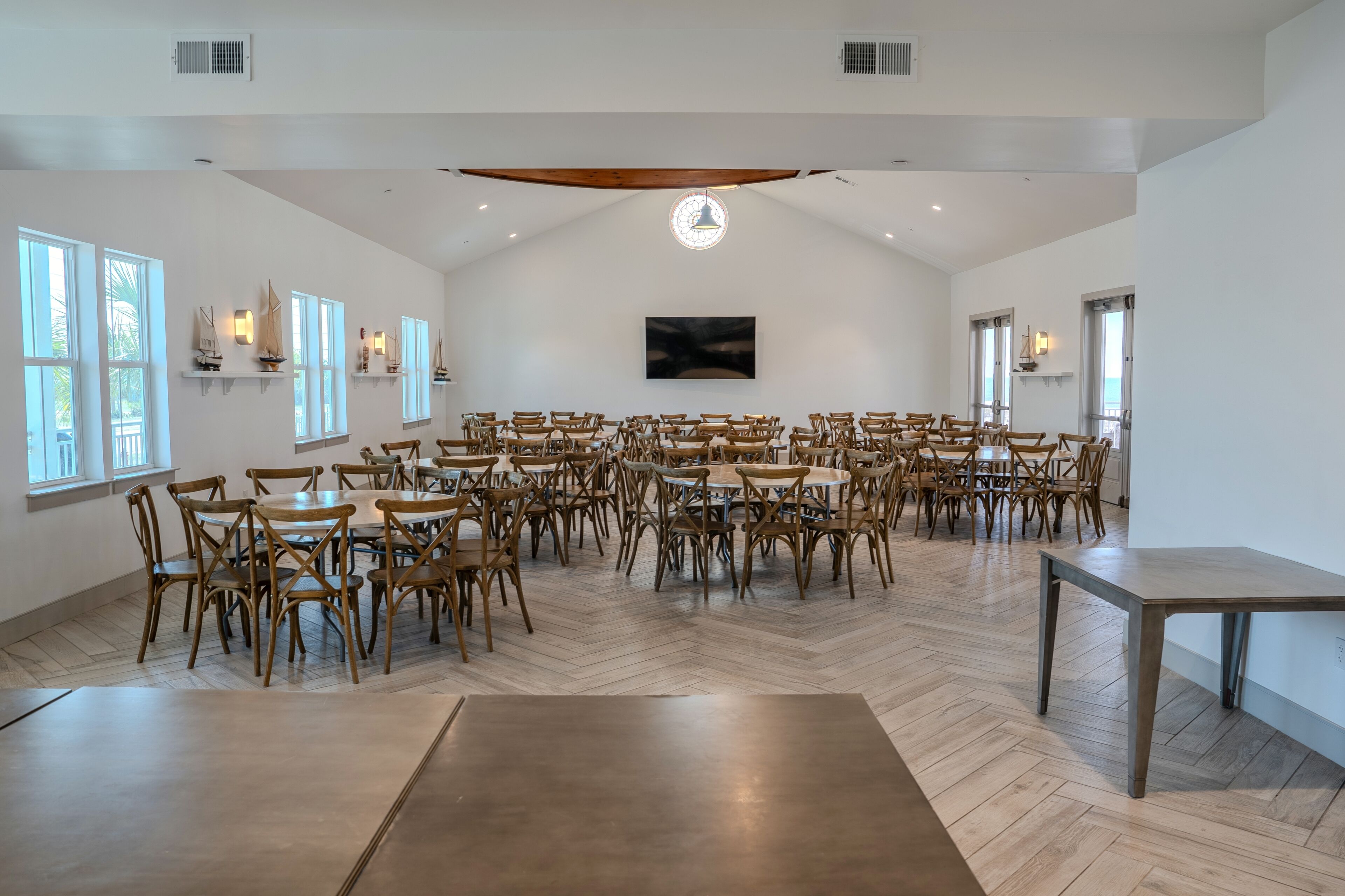 Une pièce spacieuse dotée de nombreuses tables et chaises en bois, de grandes fenêtres et d’un plafond haut avec des poutres apparentes.