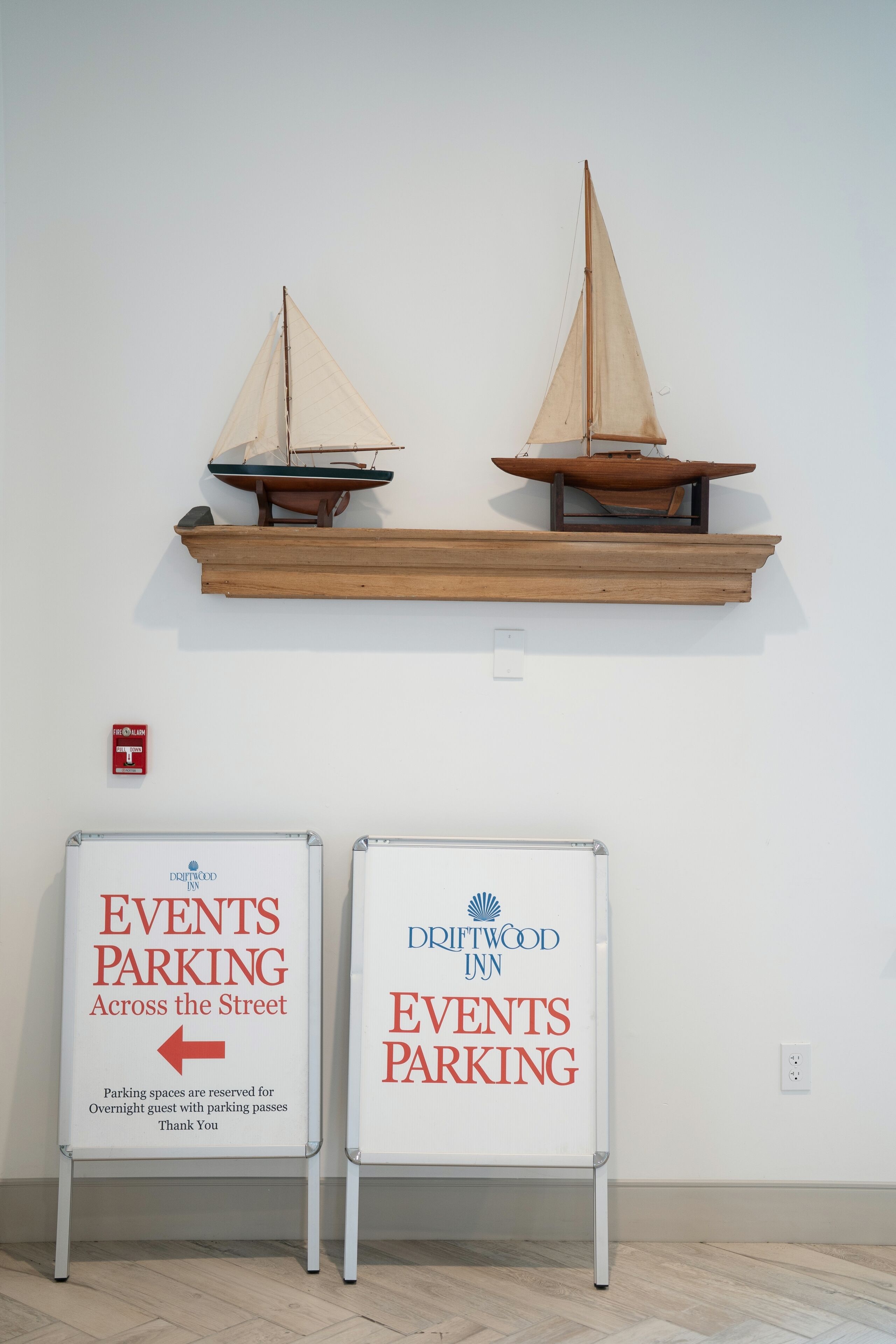 Two signs for events parking at Driftwood Inn, with a wooden shelf above displaying model sailboats.