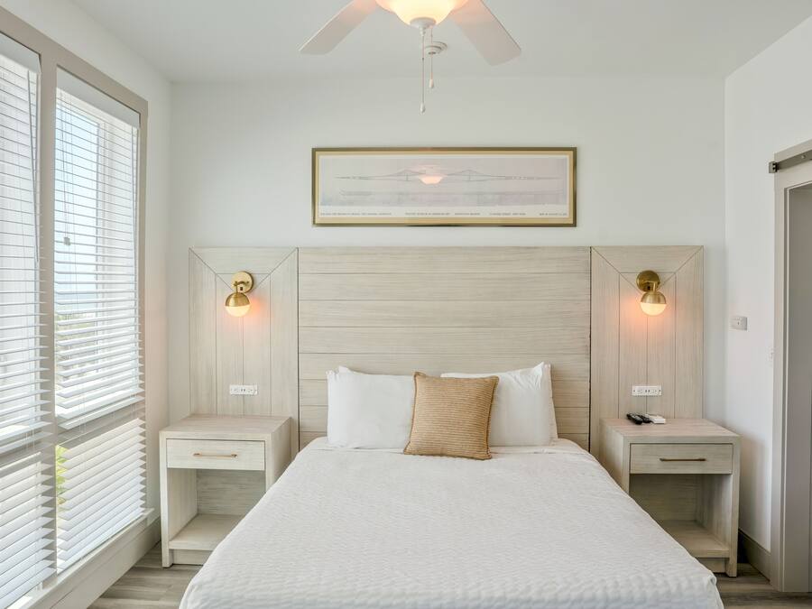 Une chambre à coucher comprenant un lit, des tables de chevet, un ventilateur de plafond, un tableau encadré et une fenêtre avec des stores.