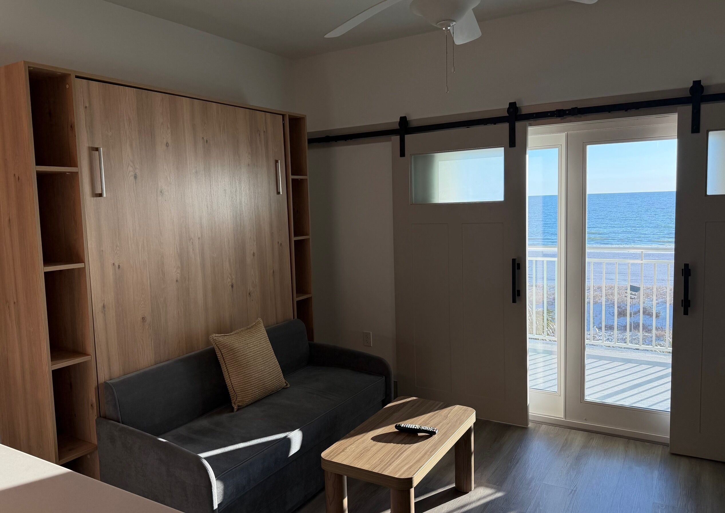 Une pièce avec un canapé gris foncé, une table en bois avec une télécommande, une armoire en bois et une porte-fenêtre coulissante donnant sur un balcon avec vue sur la mer.