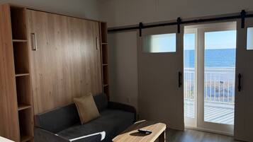 Une pièce avec un canapé gris foncé, une table en bois avec une télécommande, une armoire en bois et une porte-fenêtre coulissante donnant sur un balcon avec vue sur la mer.