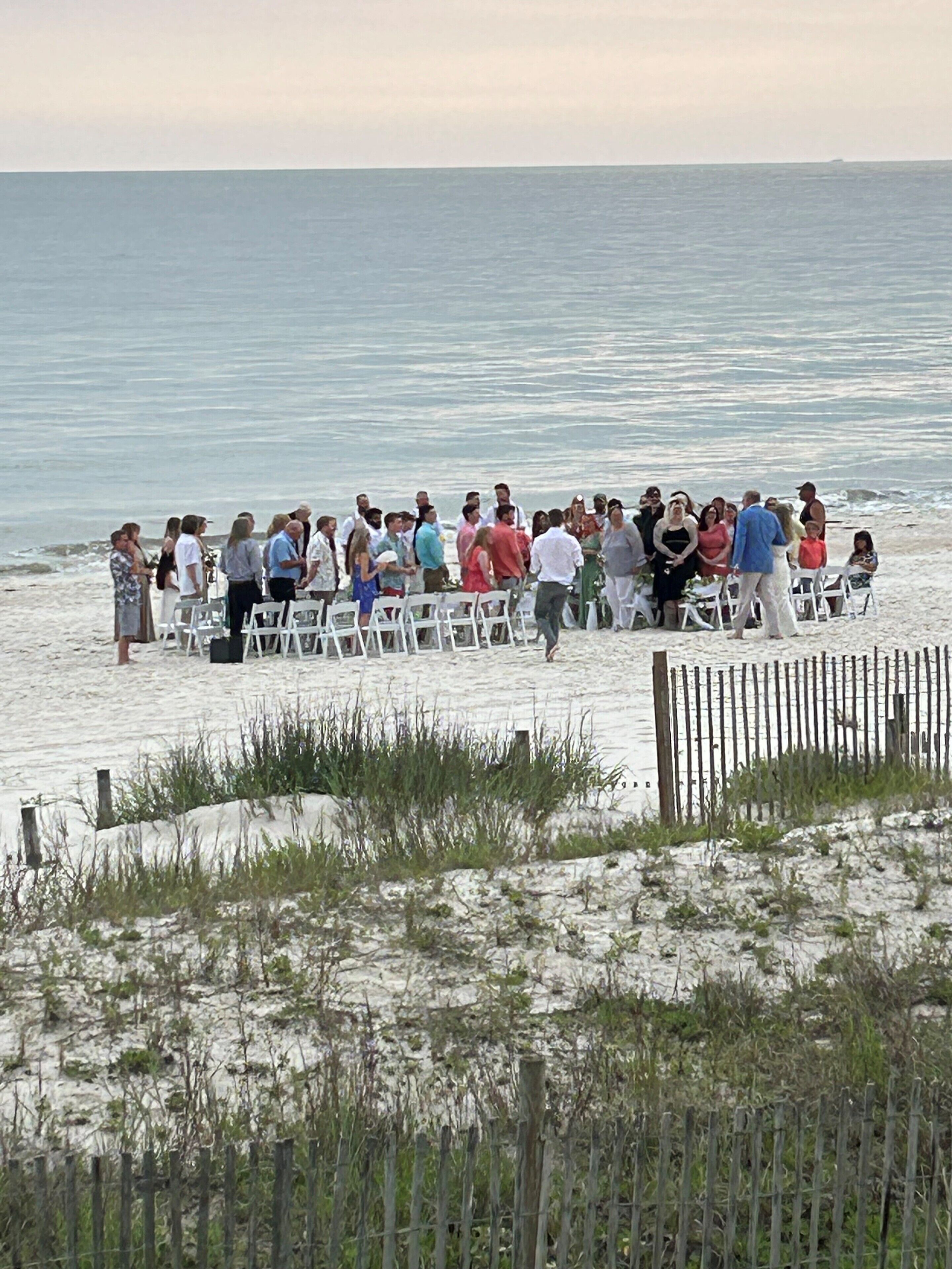 A beachside event with white chairs arranged in rows, a sandy foreground with sparse vegetation, and the ocean in the background.