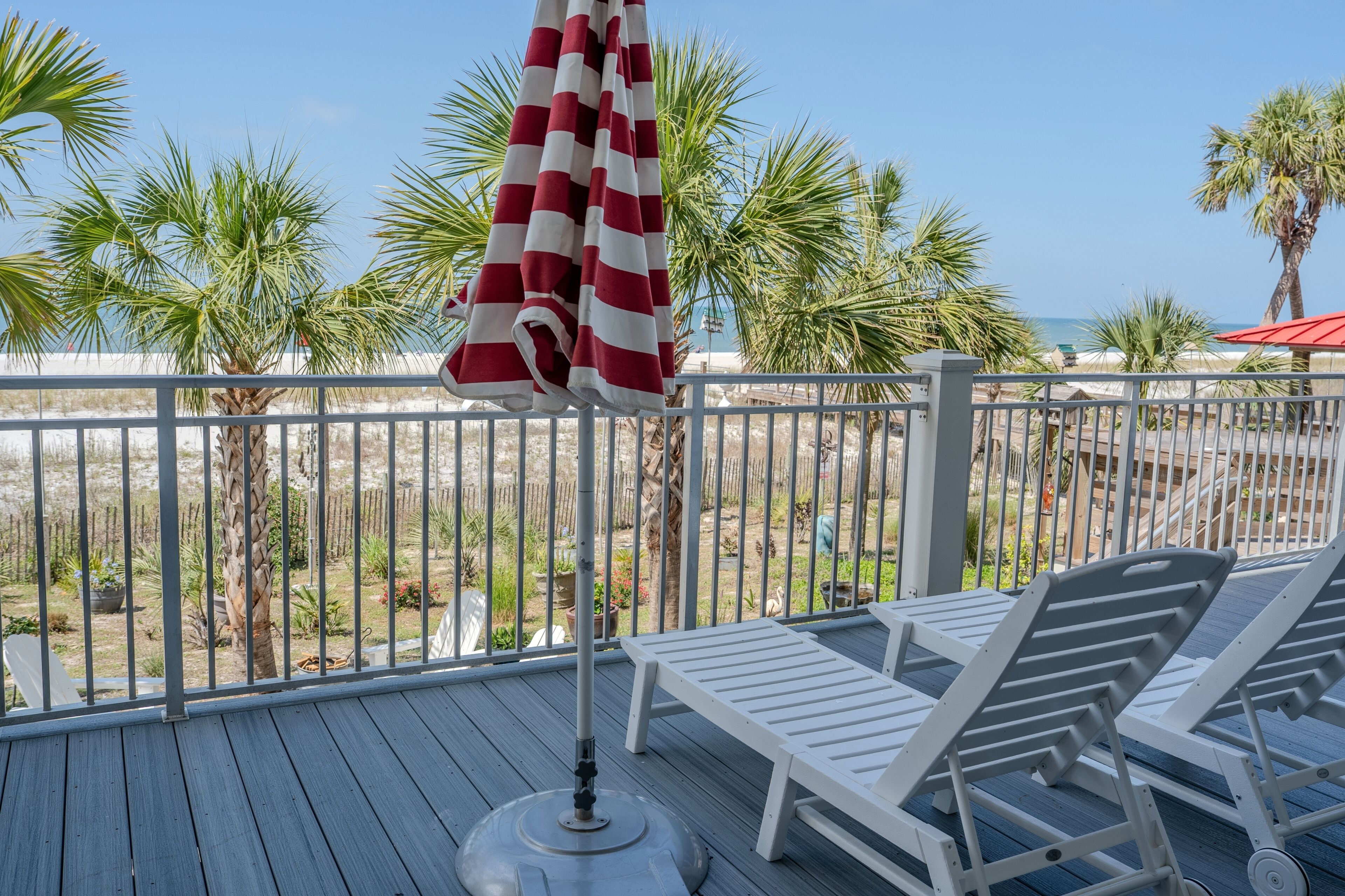 A deck with lounge chairs and a striped umbrella, overlooking a beach with palm trees and a fence.