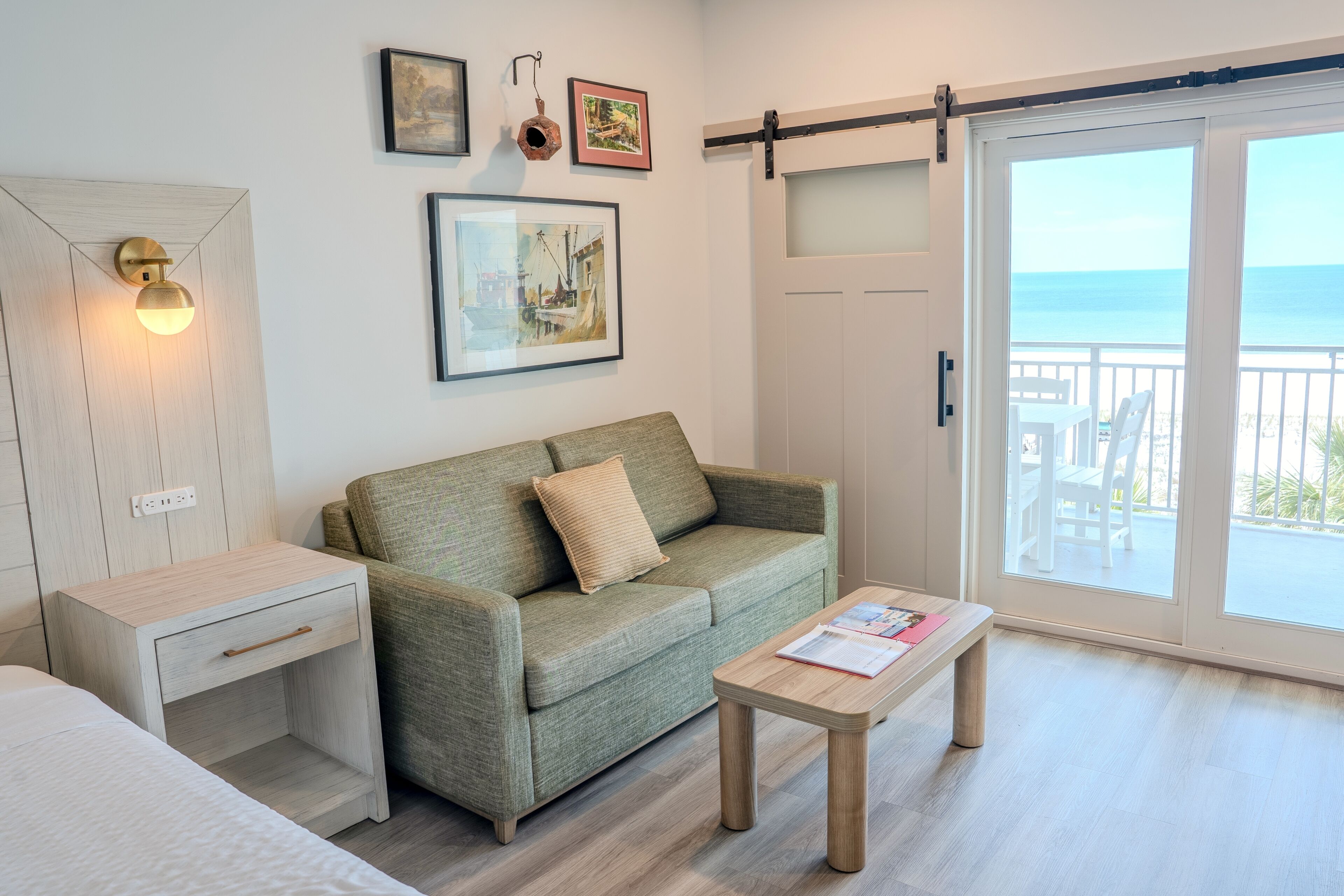 Una habitación con sofá verde, una mesita de noche de madera, una mesa de centro de madera y un ventanal corredero que da a un balcón con vistas al mar.