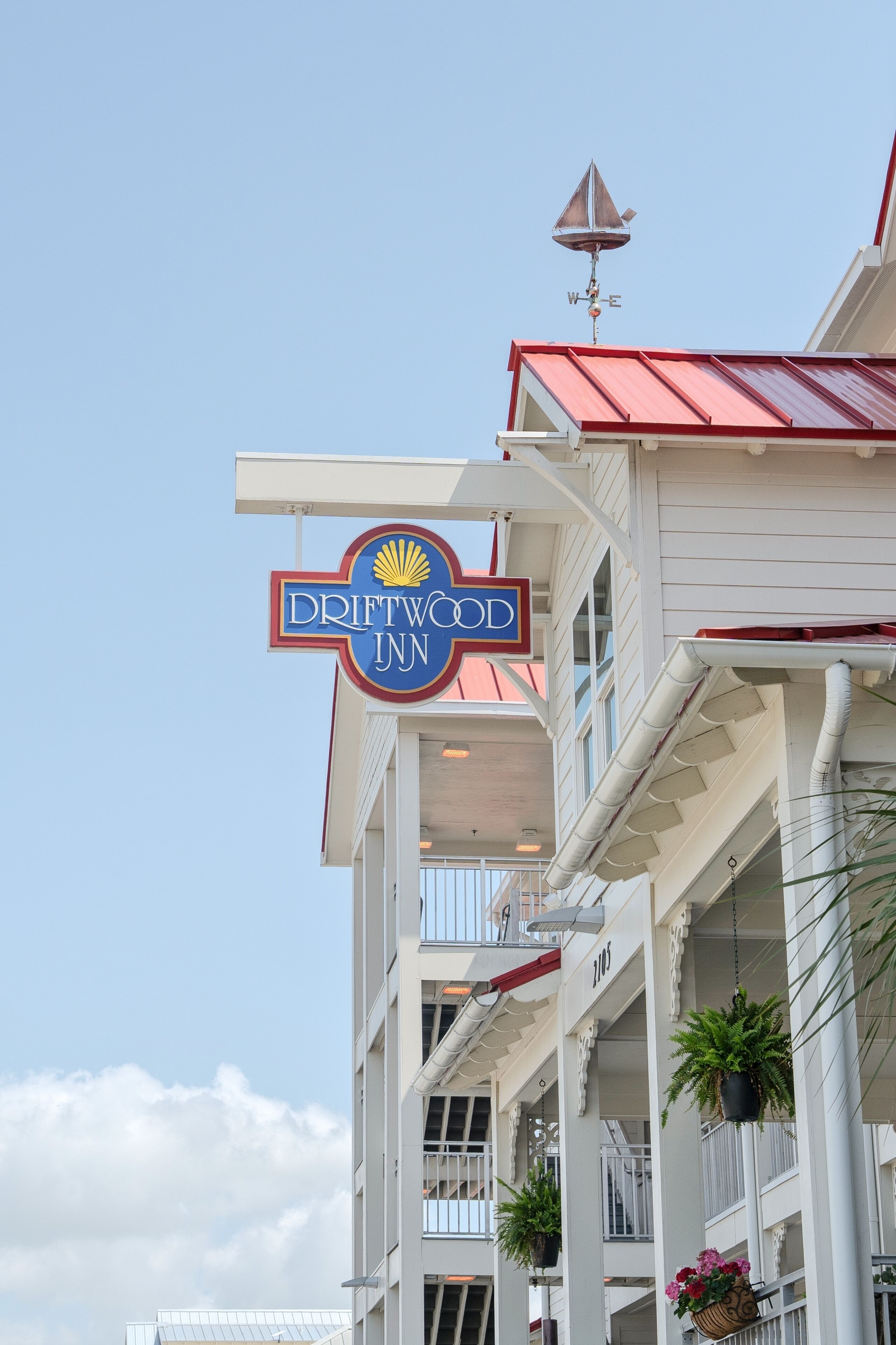 A Driftwood Inn sign with a weather vane, white building with balconies, and hanging plants.