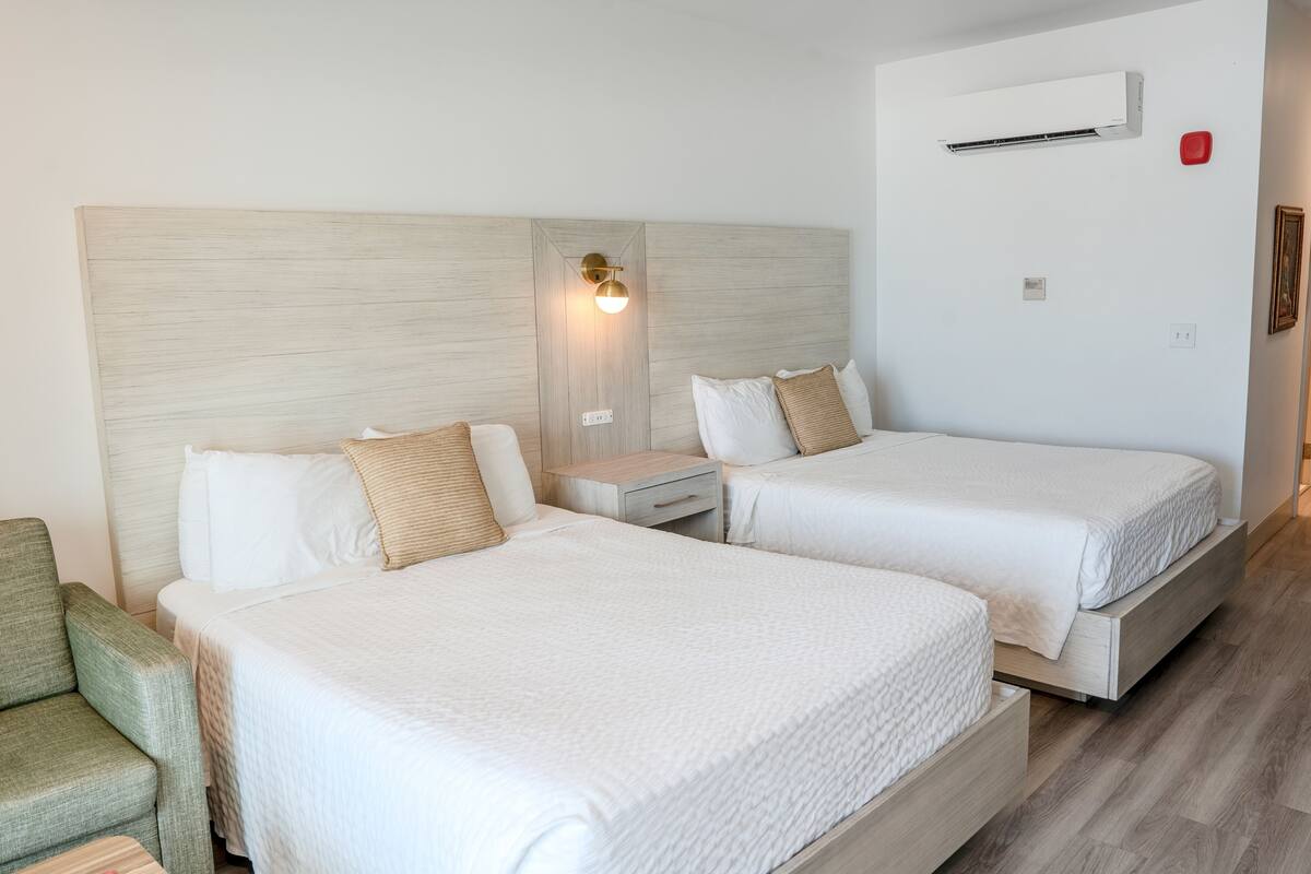 A hotel room with two beds, a nightstand, a chair, and a wall-mounted air conditioner.