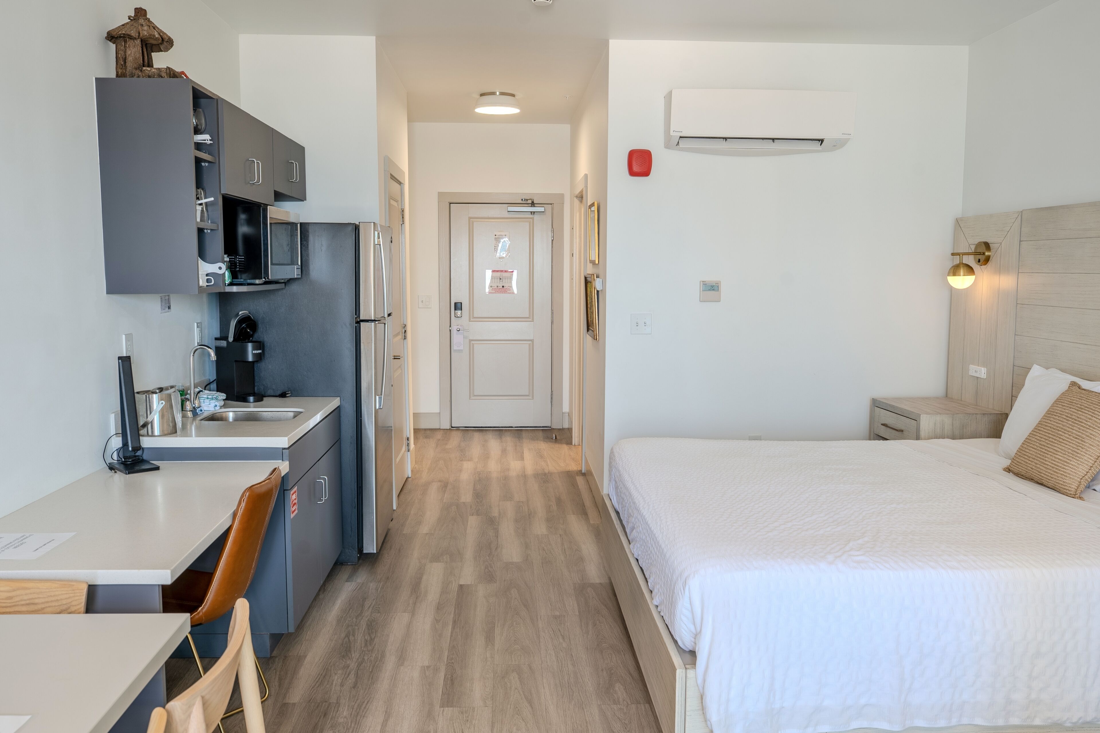 A modern hotel room with a kitchenette, a bed with a white comforter, a wooden headboard, and a wall-mounted air conditioner.