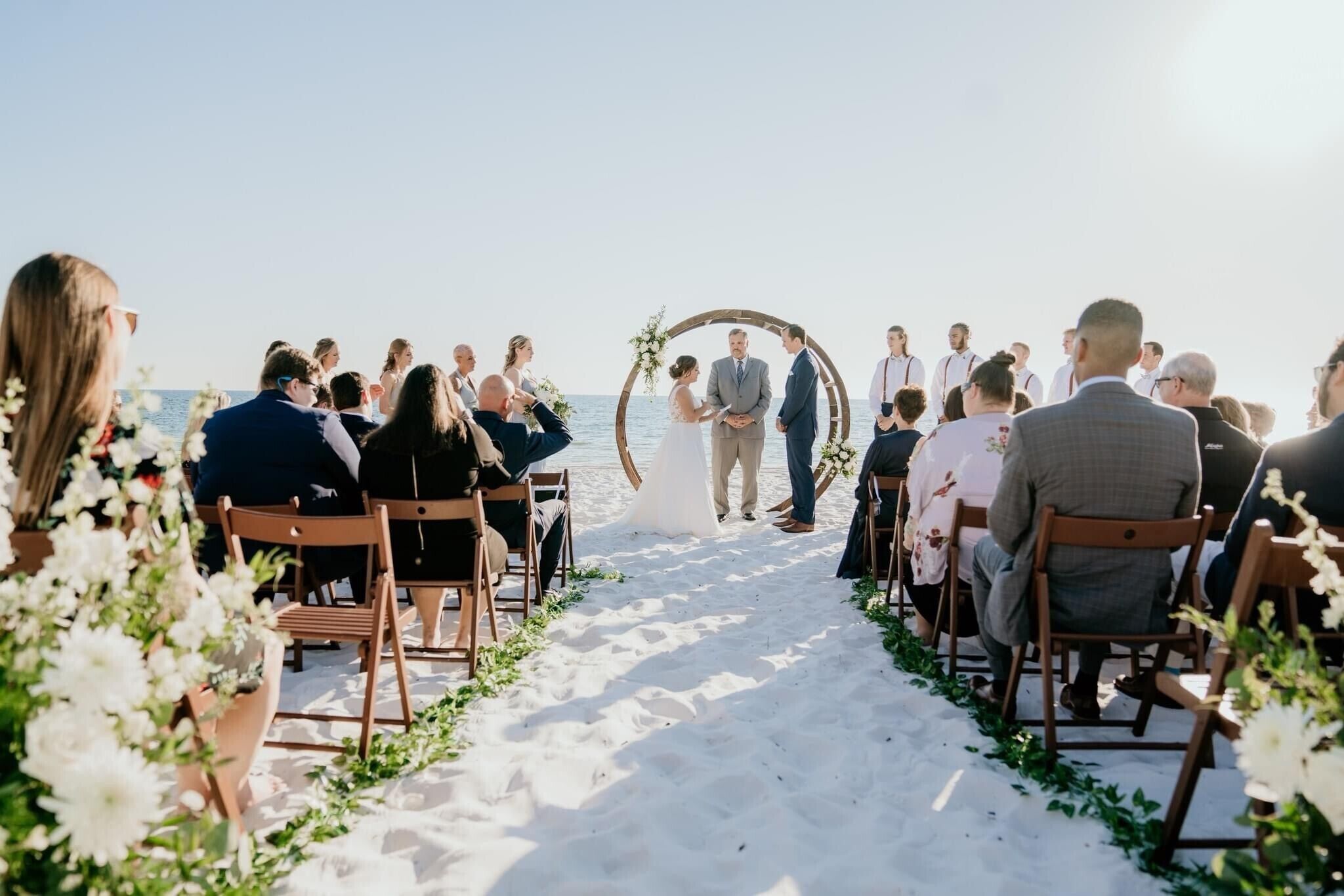 A beach wedding ceremony with guests seated on wooden chairs facing the aisle.