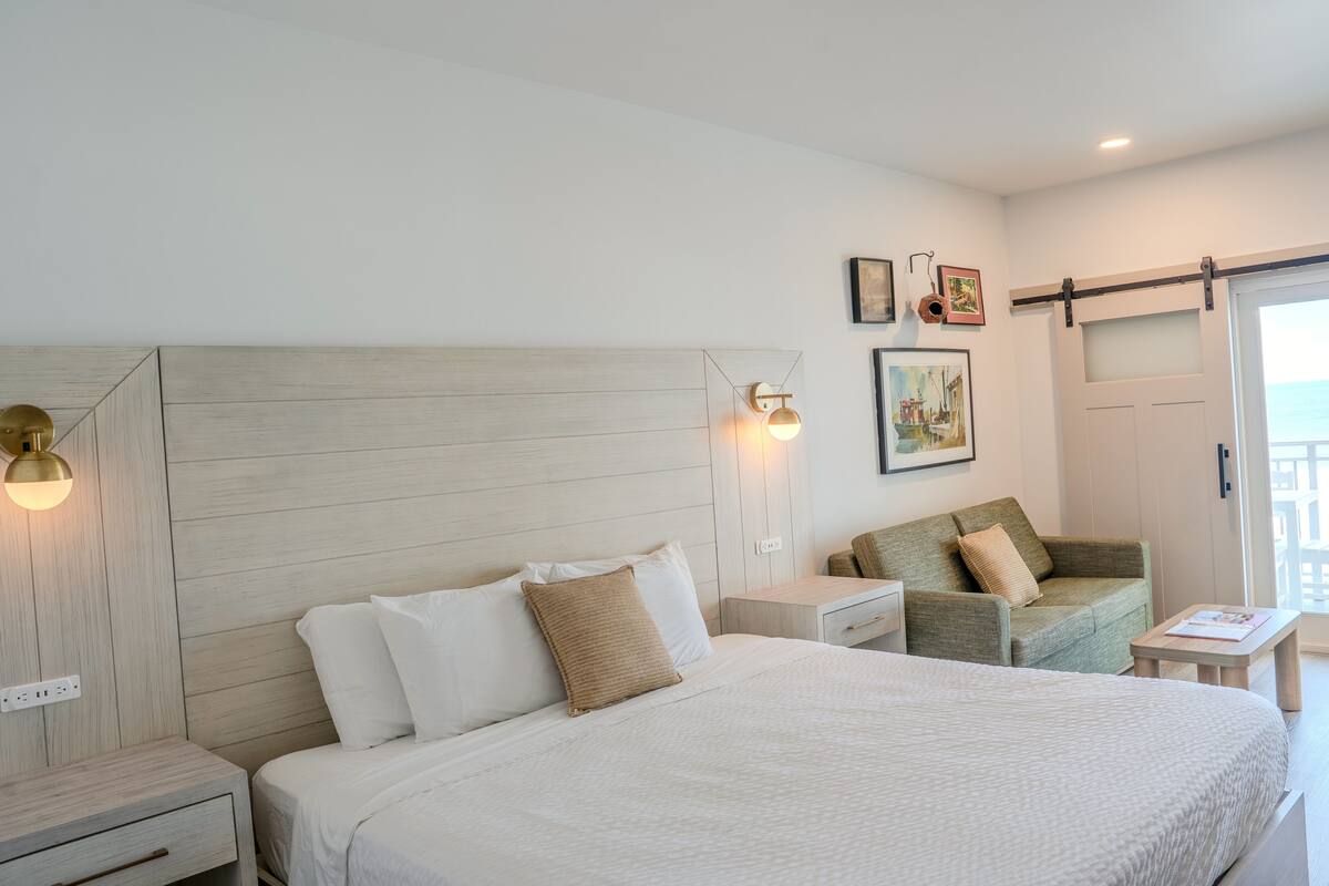 A hotel room with a large bed, a nightstand, a chair, a small table, and a sliding door to the balcony.