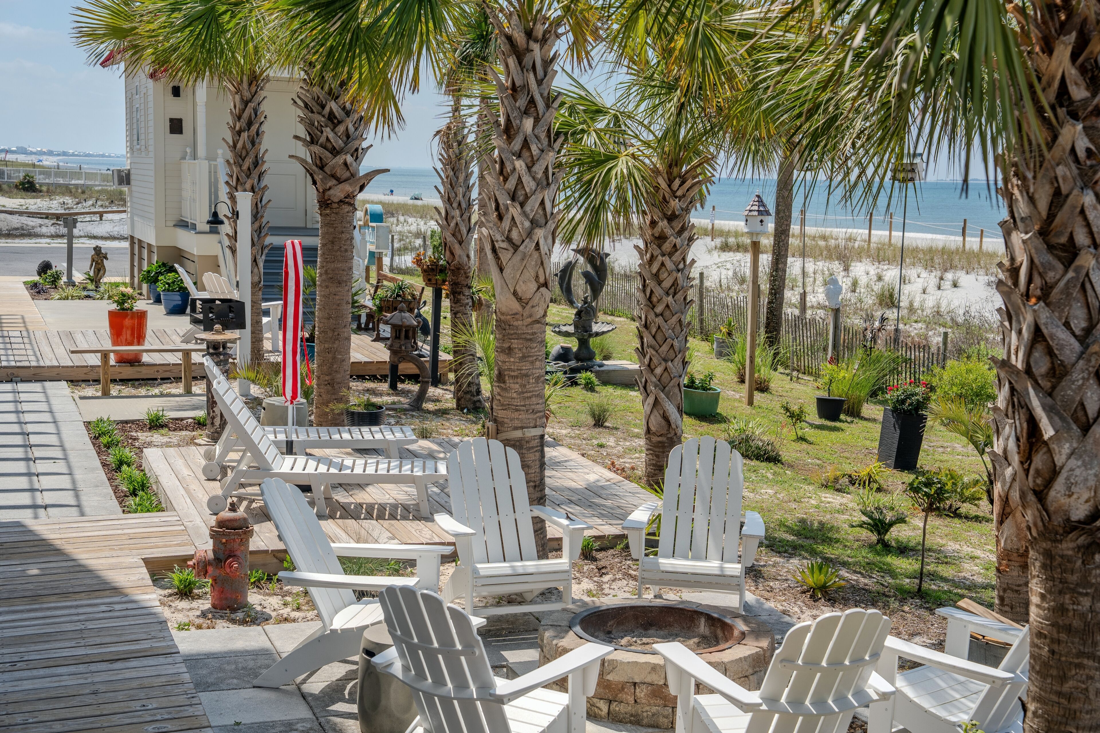 A deck with white Adirondack chairs, a fire pit, and a view of the beach.