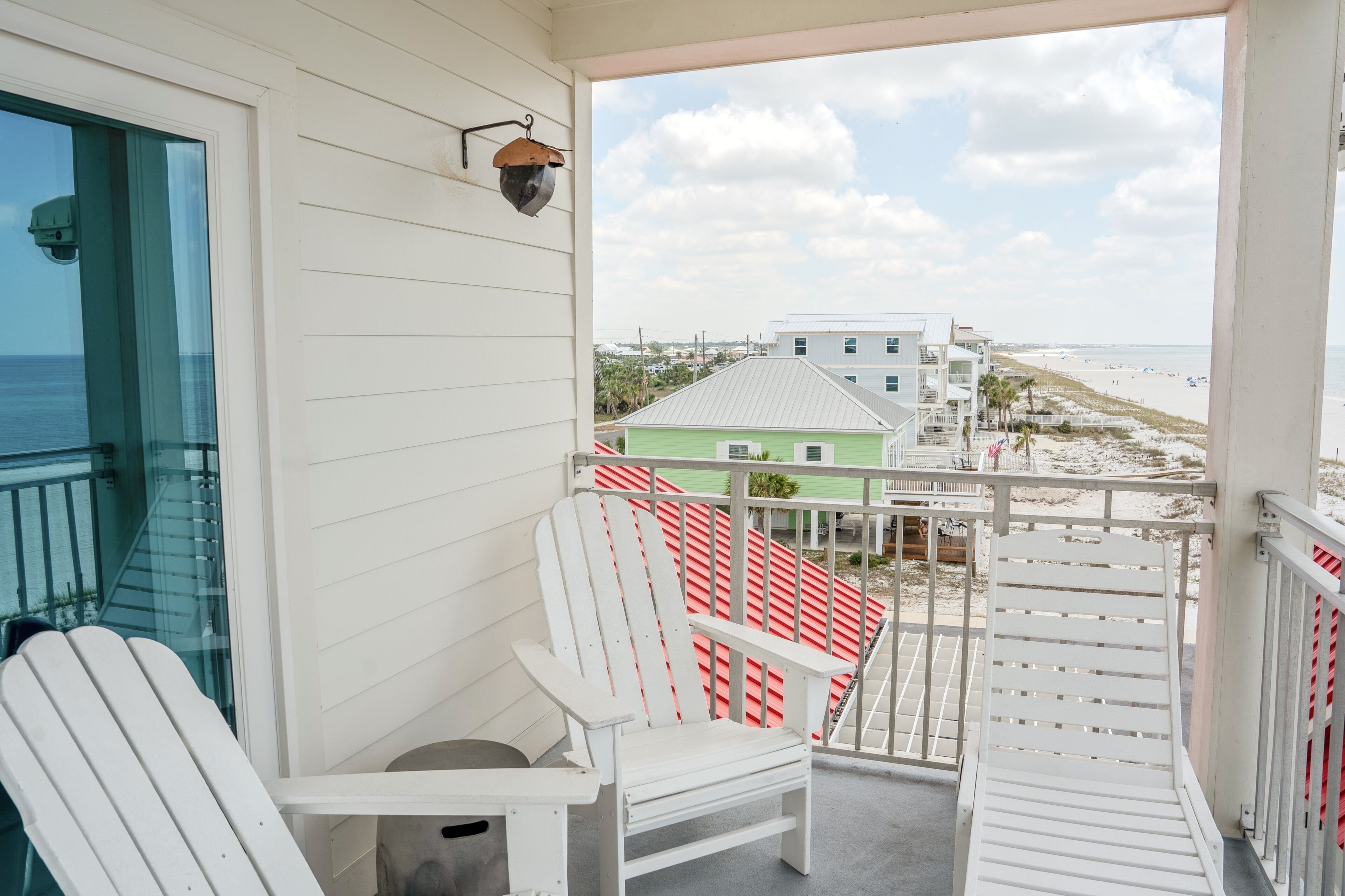 A balcony with white Adirondack chairs, a view of the beach, and a green building in the background.