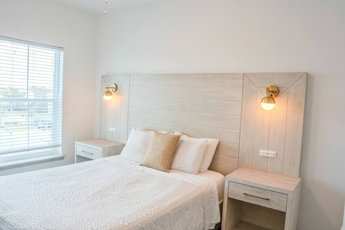 A bedroom with a large bed, two bedside tables, a nightstand, a window with blinds, and wall-mounted lighting.