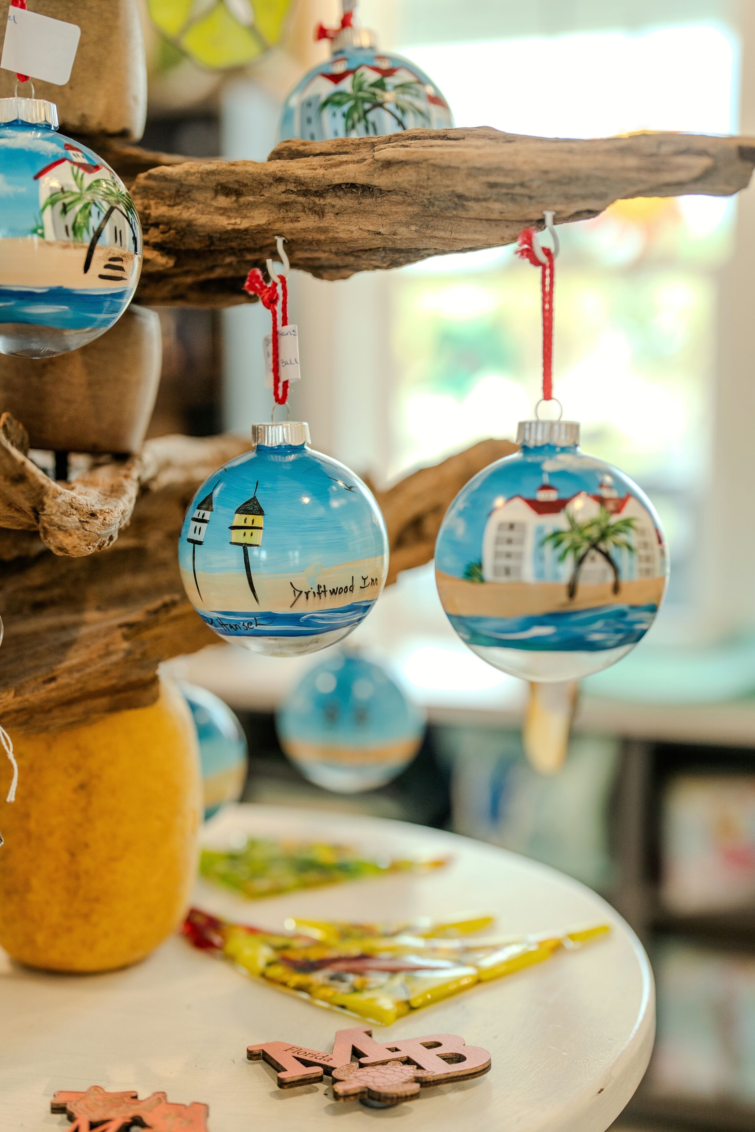A wooden branch with hanging ornaments depicting coastal scenes, a white table with scattered items, and a blurred background.