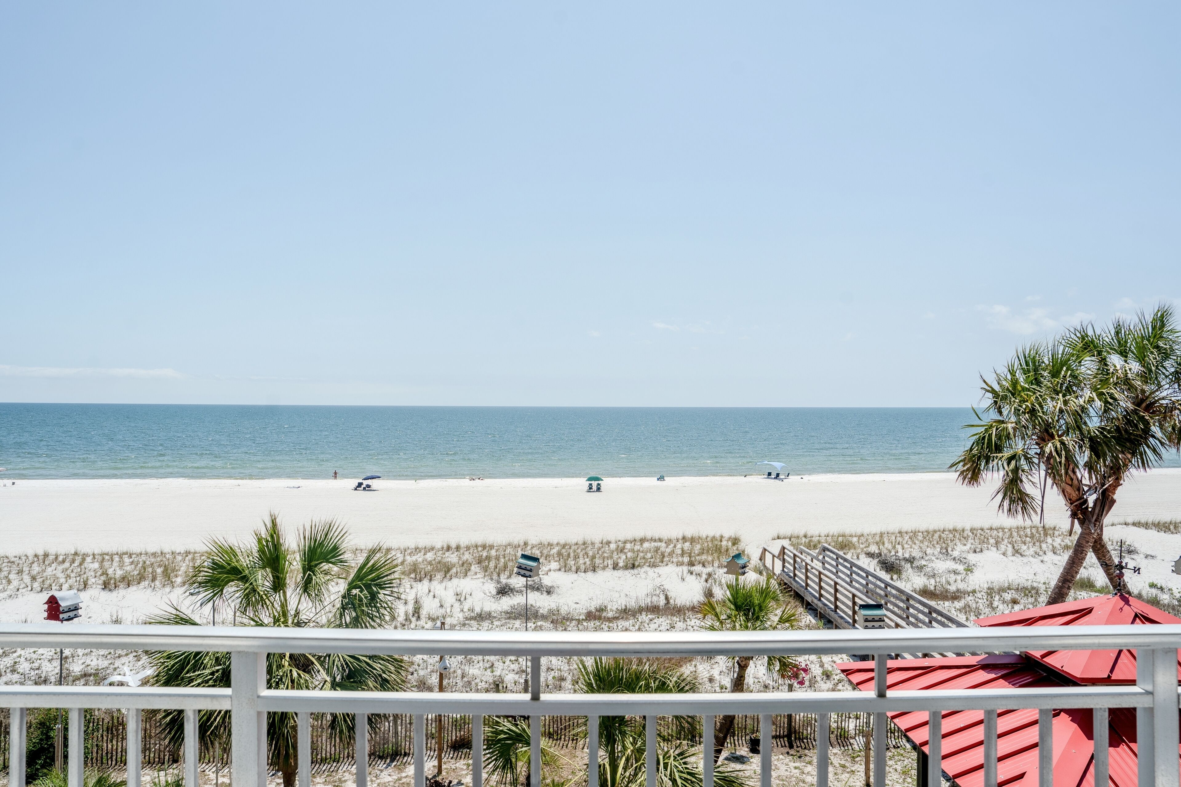 A beach view from a balcony with a railing, palm trees, and a red umbrella.