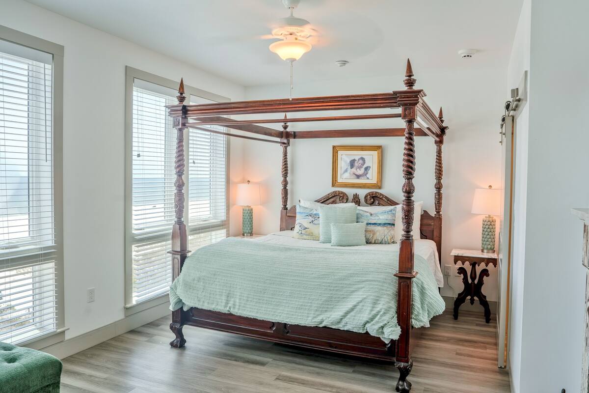 A four-poster bed with a wooden frame, a green bedspread, and matching pillows. There is a nightstand with a lamp, a framed picture on the wall, and a small table with a vase.