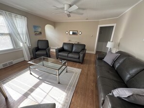 A living room with a glass coffee table, two leather sofas, a ceiling fan, and a window with curtains. - Charming Southold Retreat with In Ground Pool – Walk to Founder’s Beach (Southold)