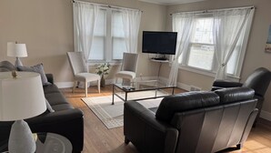 Un salon moderne avec un canapé en cuir noir, une table basse en verre et un téléviseur à écran plat. On y trouve deux rideaux blancs, un vase avec des fleurs et une lampe sur une table d’appoint.