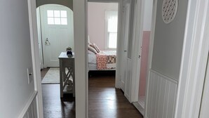 Un couloir au plancher de bois et aux murs blancs, qui mène à une chambre à coucher avec un lit et une fenêtre.
