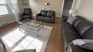 Un salon avec une table basse en verre, deux canapés en cuir noir, un ventilateur de plafond et une fenêtre avec des rideaux blancs.