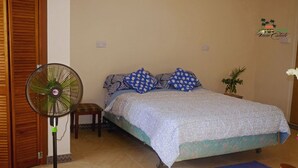 A bedroom with a bed, a standing fan, a small stool, a potted plant, and a wooden door.