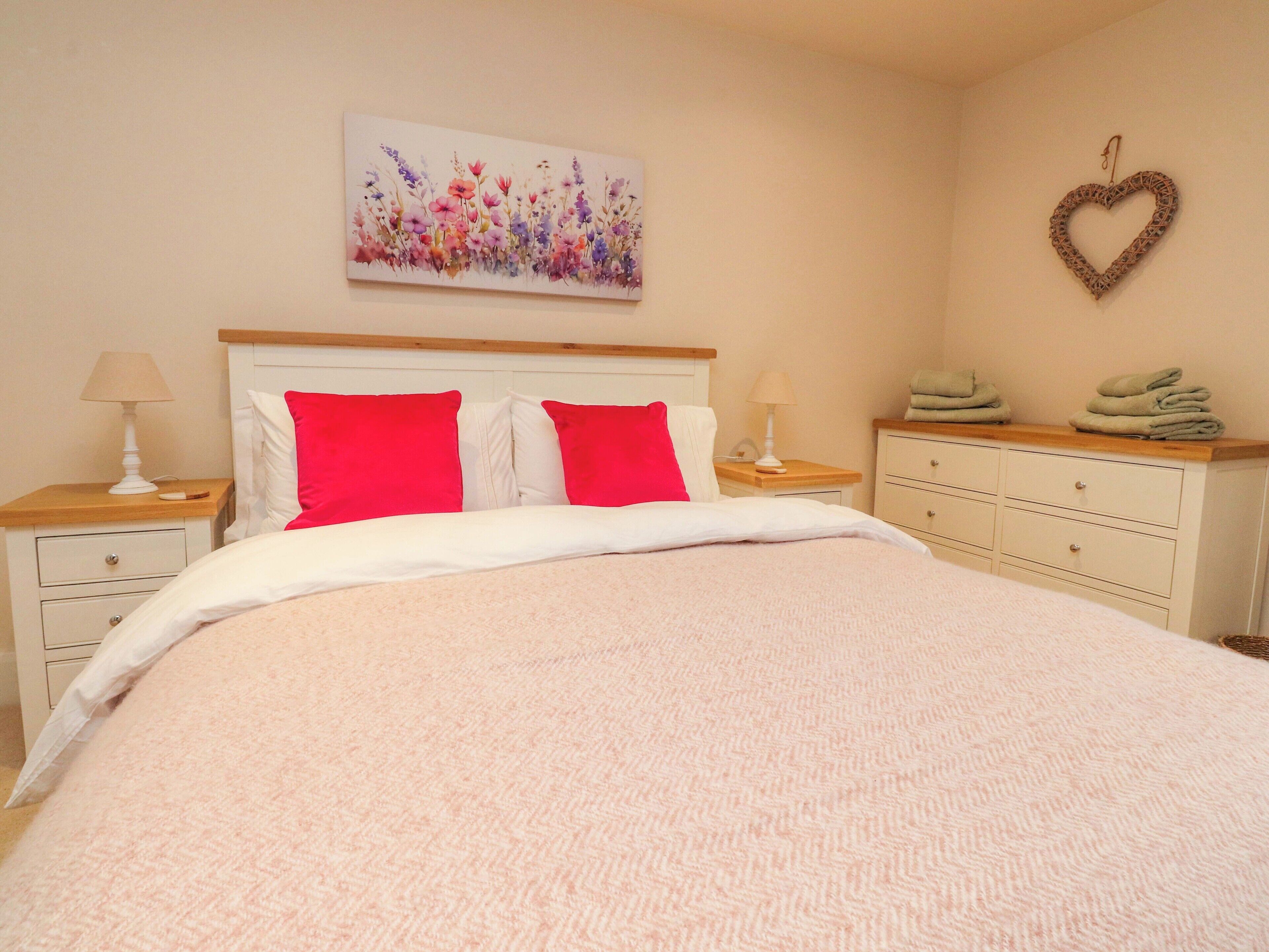 A neatly made bed with a pink blanket, two red pillows, and a floral artwork above it.