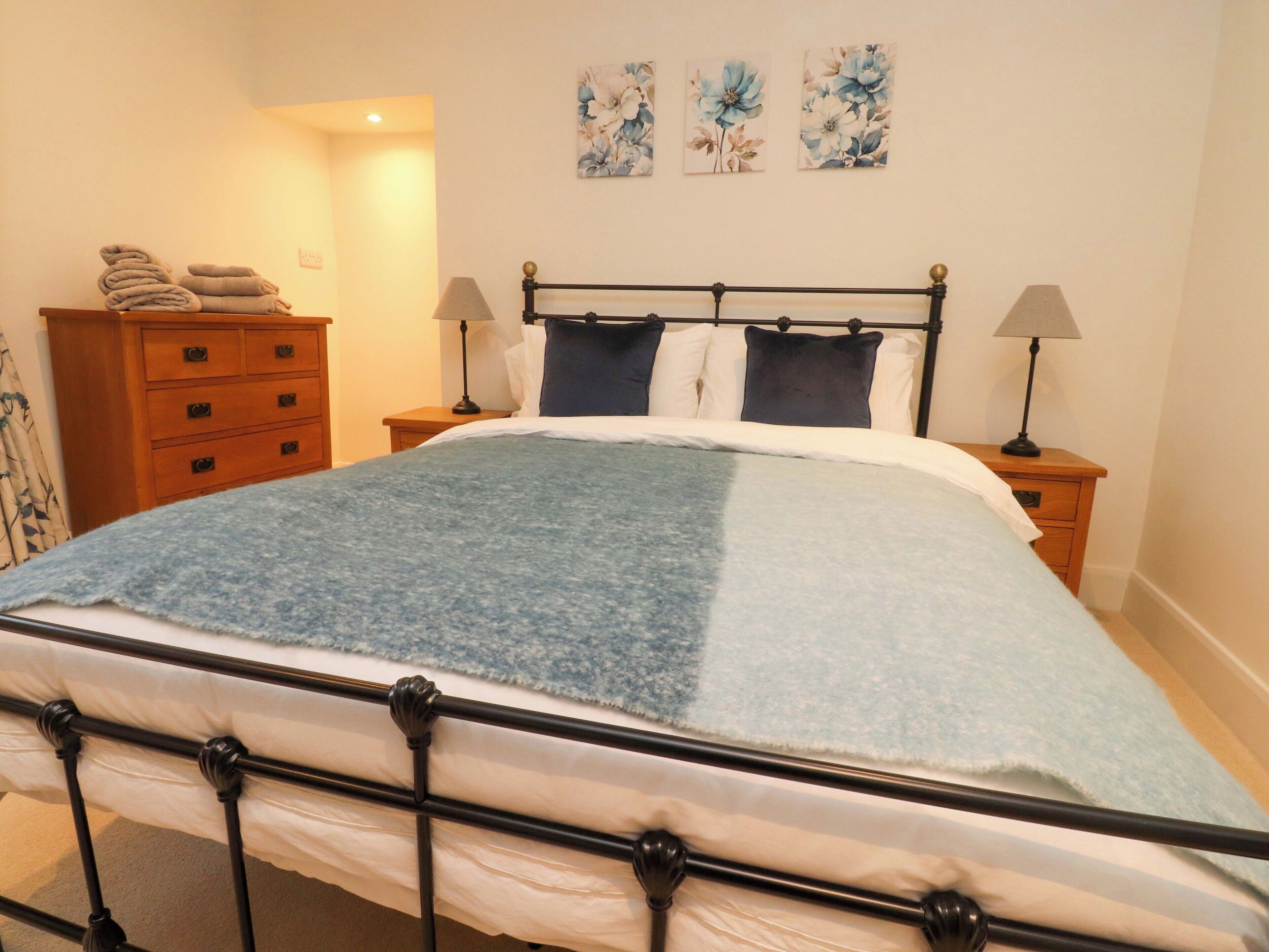A neatly made bed with a blue and white bedspread, flanked by two bedside tables with lamps, a wooden dresser, and floral wall art.