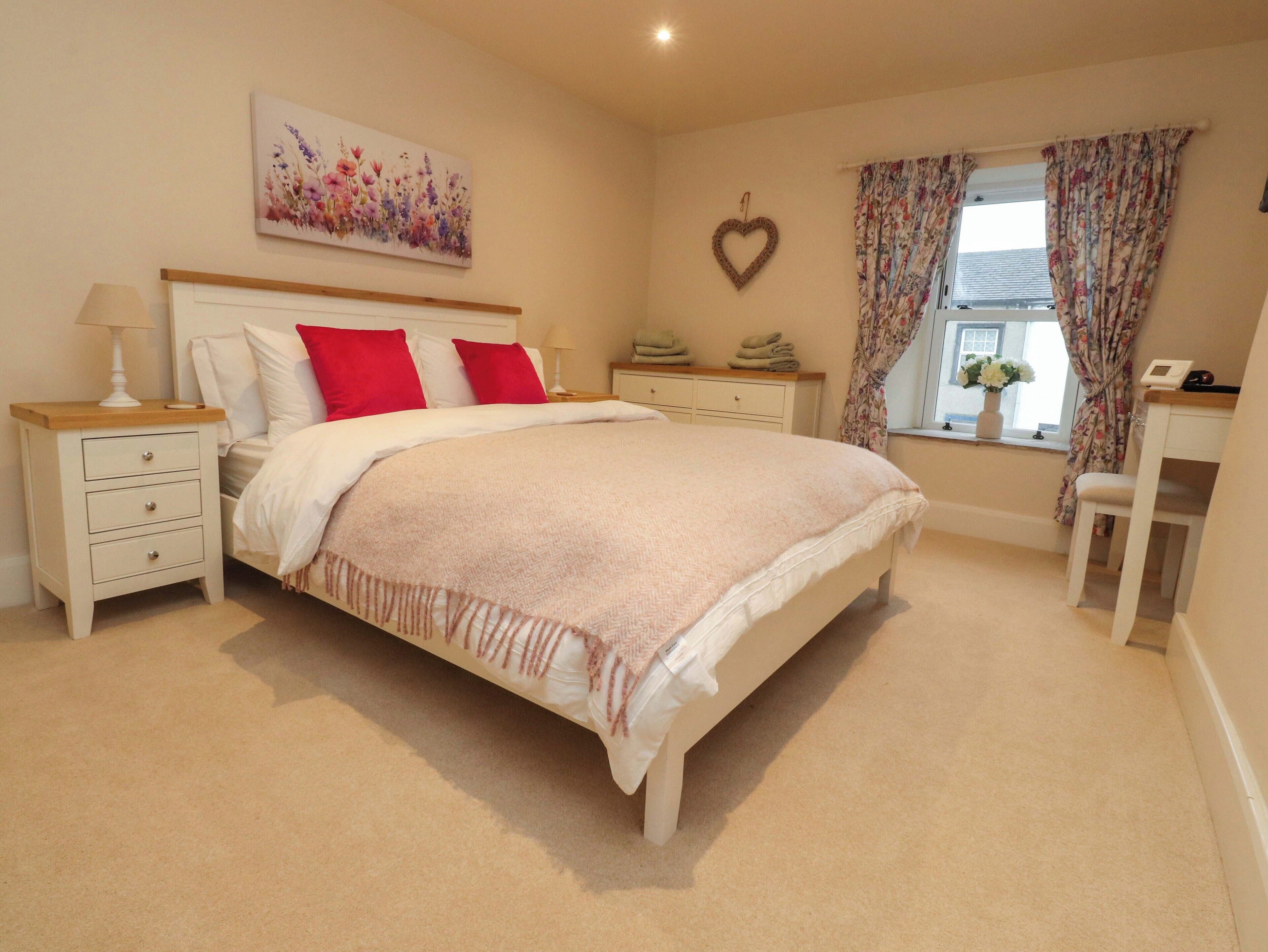 A bedroom with a large bed, two bedside tables, a nightstand, a dresser, a window with curtains, and a wall-mounted heart decoration.