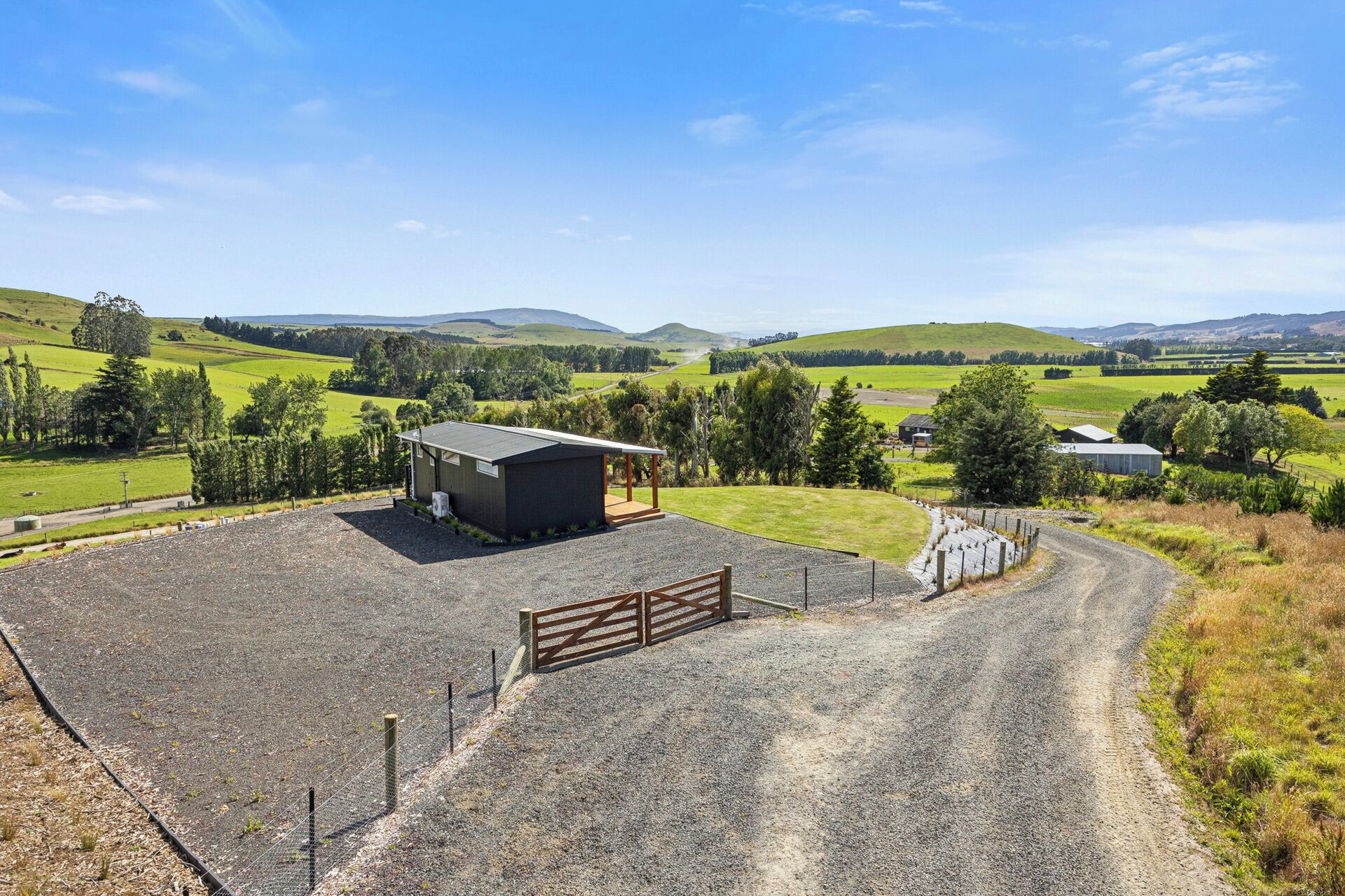 A gravel driveway leading to a fenced area with a small building, surrounded by green fields and hills.