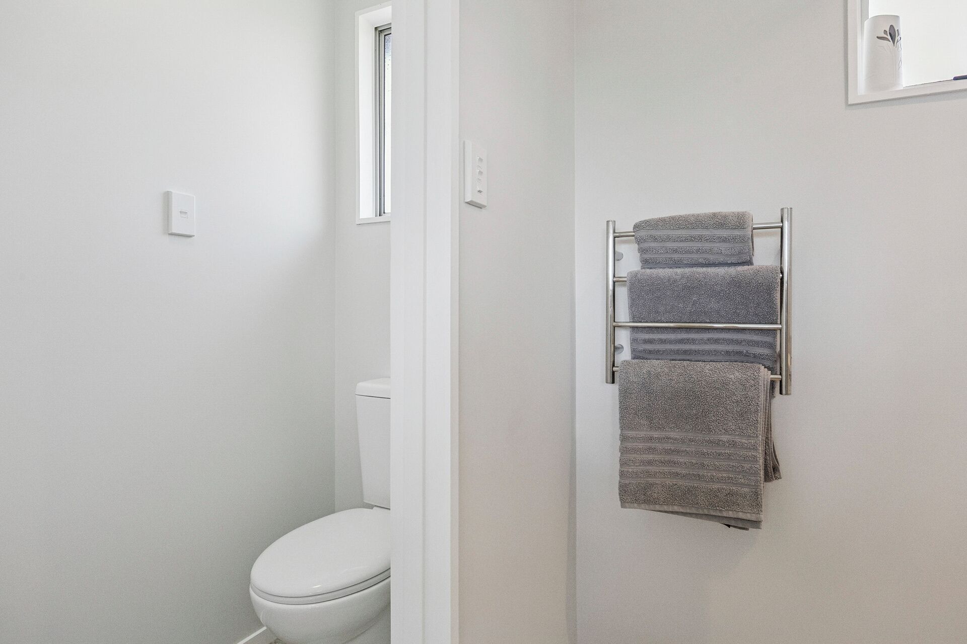 A bathroom with a white toilet, a towel rack with folded towels, and a window above the toilet.