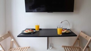 Ein Zimmer mit an der Wand befestigtem Fernseher, einem Tisch mit Frühstücksangeboten, zwei Stühlen und einer Steckdose.