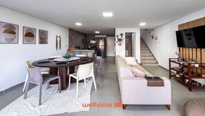 A modern living space with a dining area, a sofa, a TV, and a staircase leading to an upper level. - Triplex em Porto de Galinhas a 50m da Praia Maracaipe (Ipojuca)