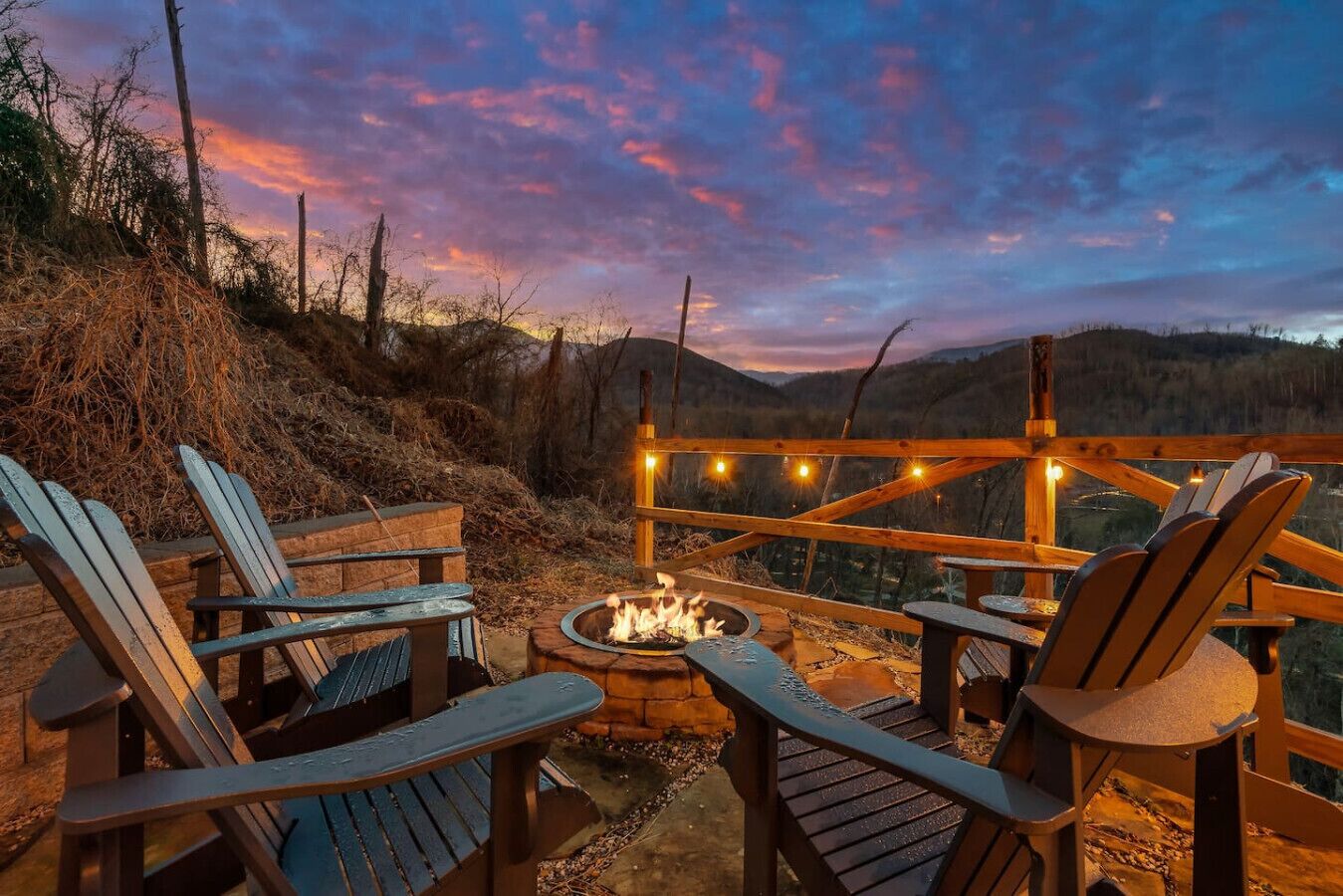 Um deck de madeira com cadeiras Adirondack e um braseiro, com vista para uma paisagem montanhosa.