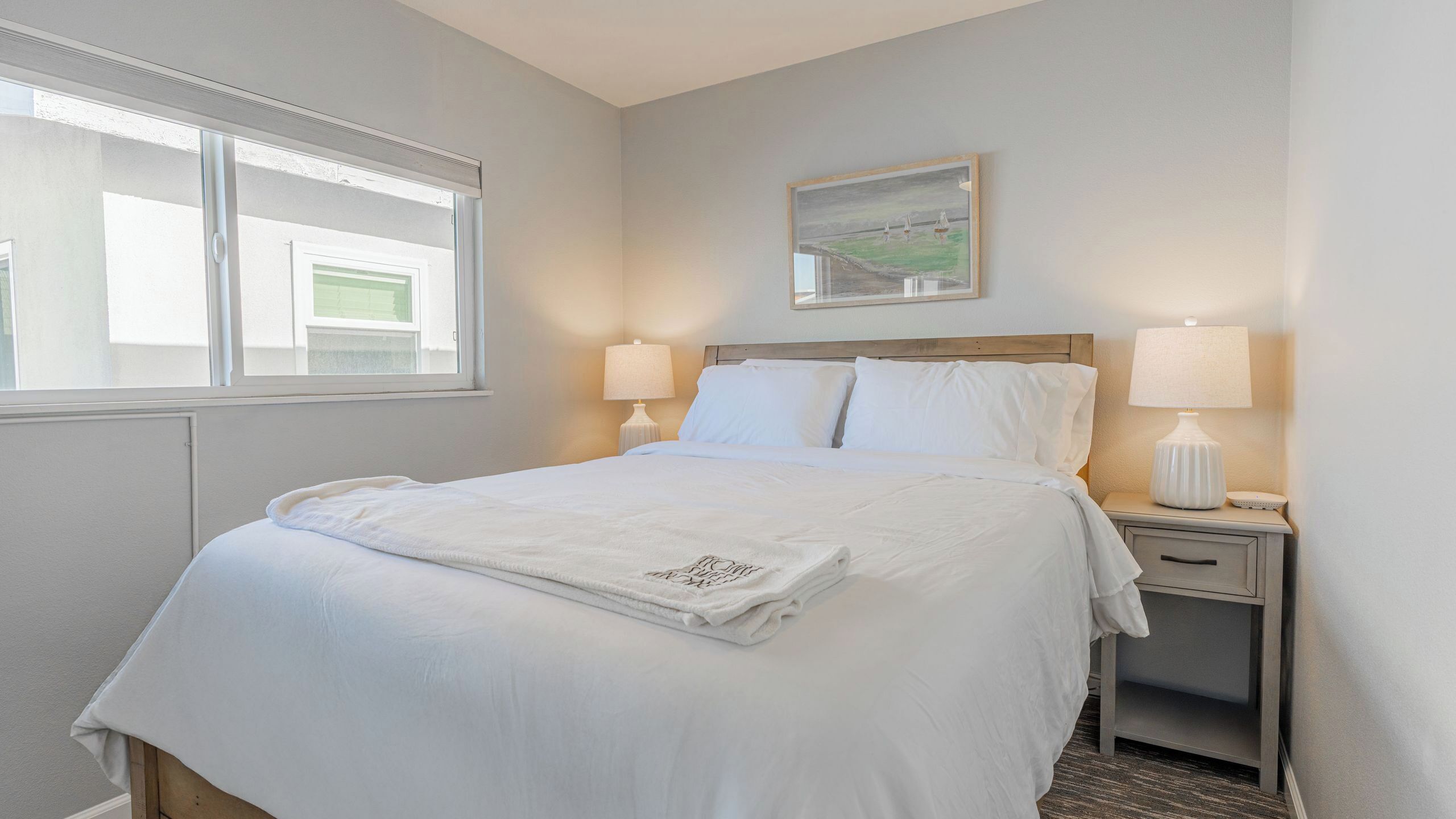 A neatly made bed with white linens, a bedside table with a lamp, and a painting on the wall.