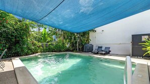Una piscina con un telo ombreggiante blu, circondata dal verde e da un muro bianco.