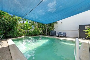 Ein Swimmingpool unter einem blauen Sonnensegel, umgeben von Grünpflanzen und einer weißen Mauer.