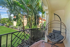 A balcony with a hanging chair, a view of a green lawn, and a building in the background.