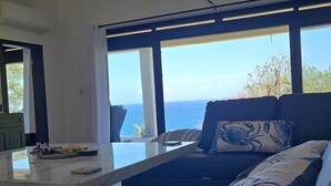 Een moderne woonkamer met een groot raam met uitzicht op de oceaan, een bank met decoratieve kussens, een tafel met een afstandsbediening en een plafondventilator.