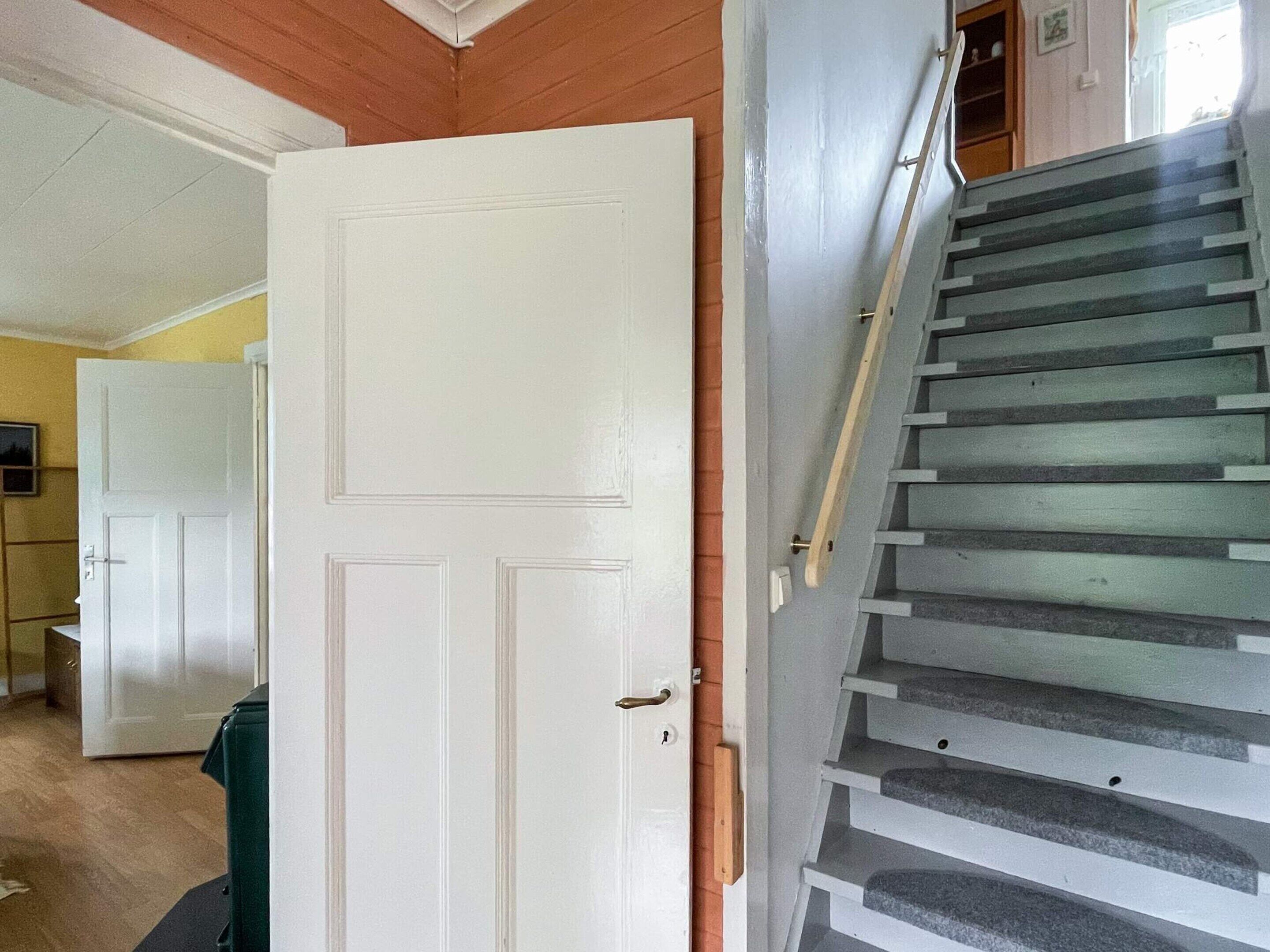 Uma escada com corrimão leva a um patamar, uma porta branca e um corredor com piso de madeira.