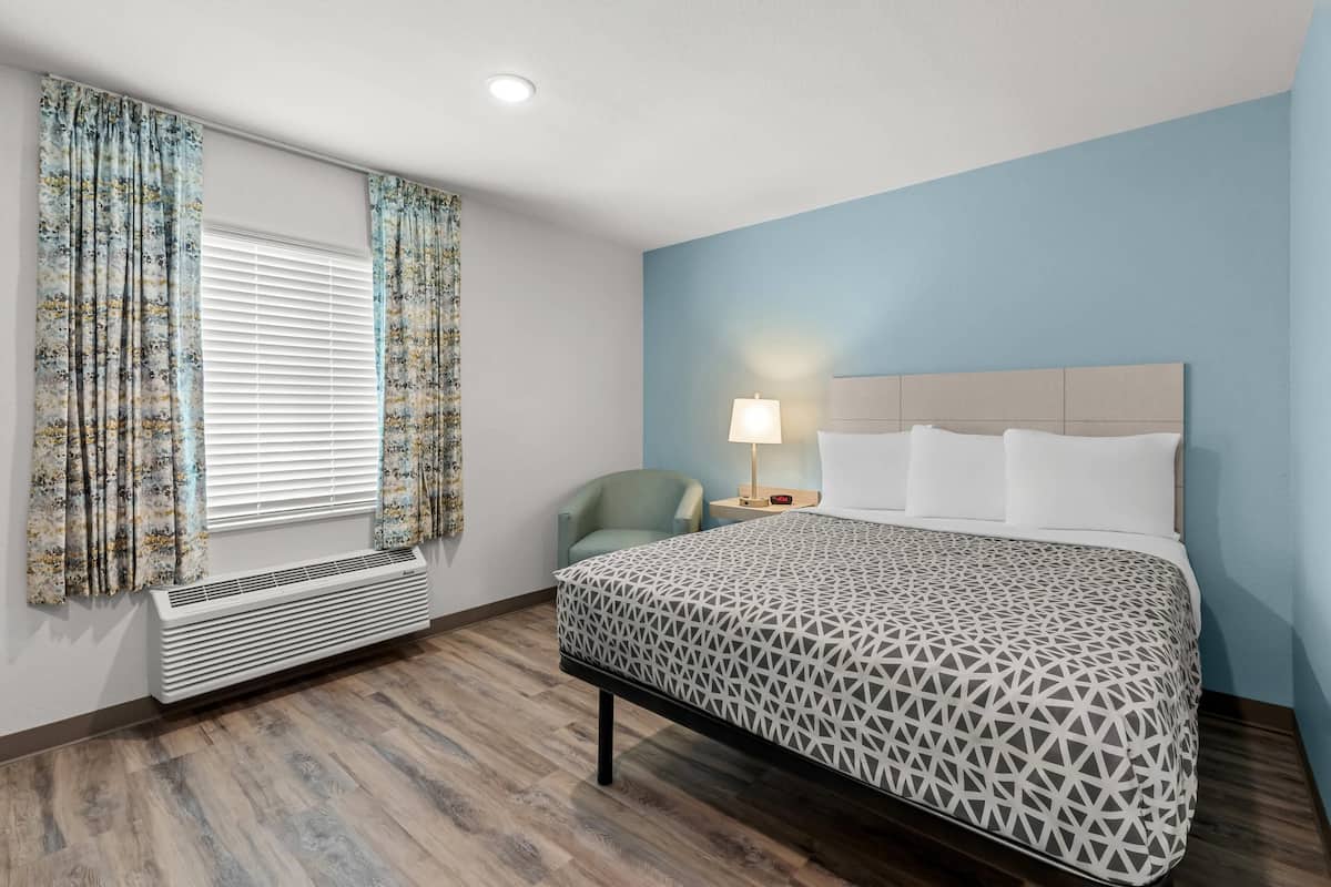Habitación de hotel con cama, una silla, mesita de noche con lámpara y ventana con cortinas.