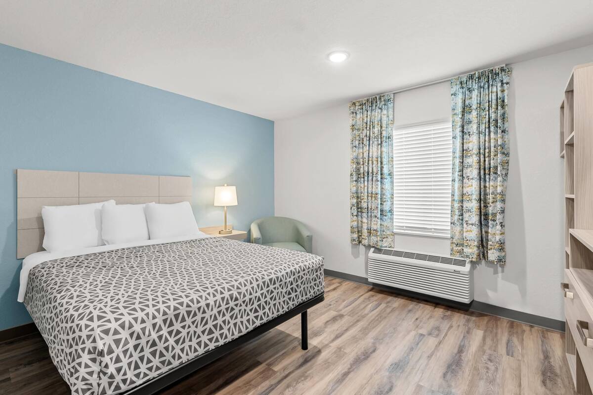 Una habitación de hotel con cama, una silla, mesita de noche con lámpara, ventana con cortinas y aire acondicionado integrado.